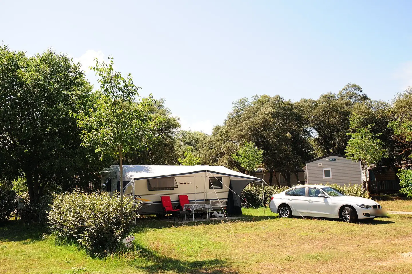 Caravane avec auvent sur un emplacement ombragé, voiture immatriculée aux Pays-Bas garée à côté, mobile-home en arrière-plan