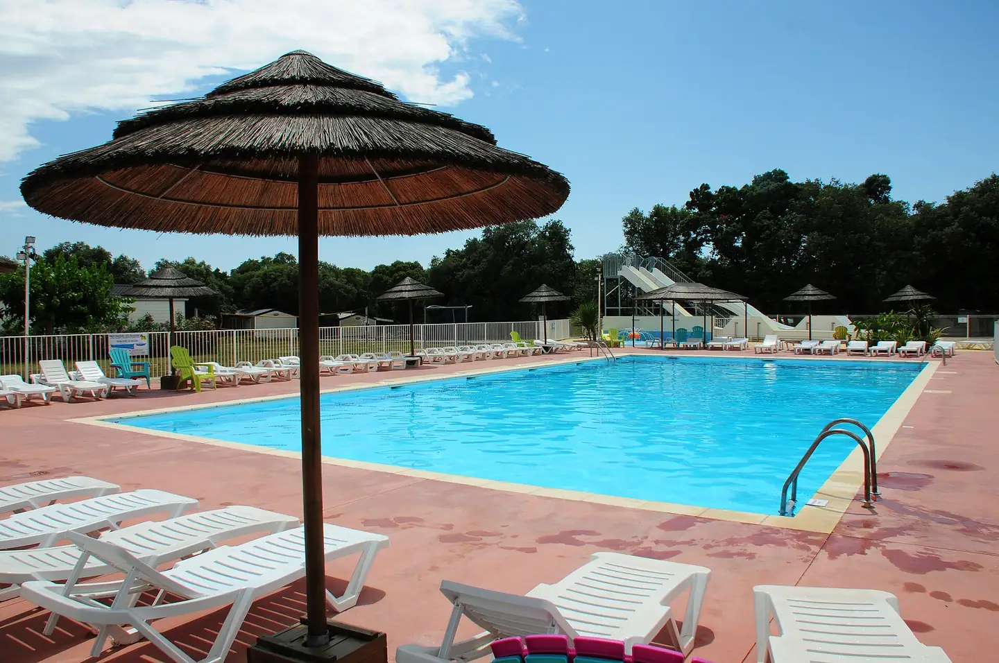 Piscine extérieure d'un camping du sud de la France, transats blancs, paillotes et toboggans en arrière-plan