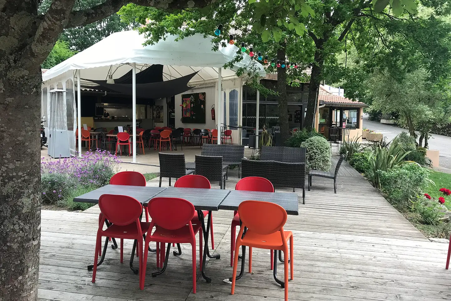 Terrasse en bois du snack-bar d'un camping, tables et chaises rouges sous les arbres, guirlande lumineuse